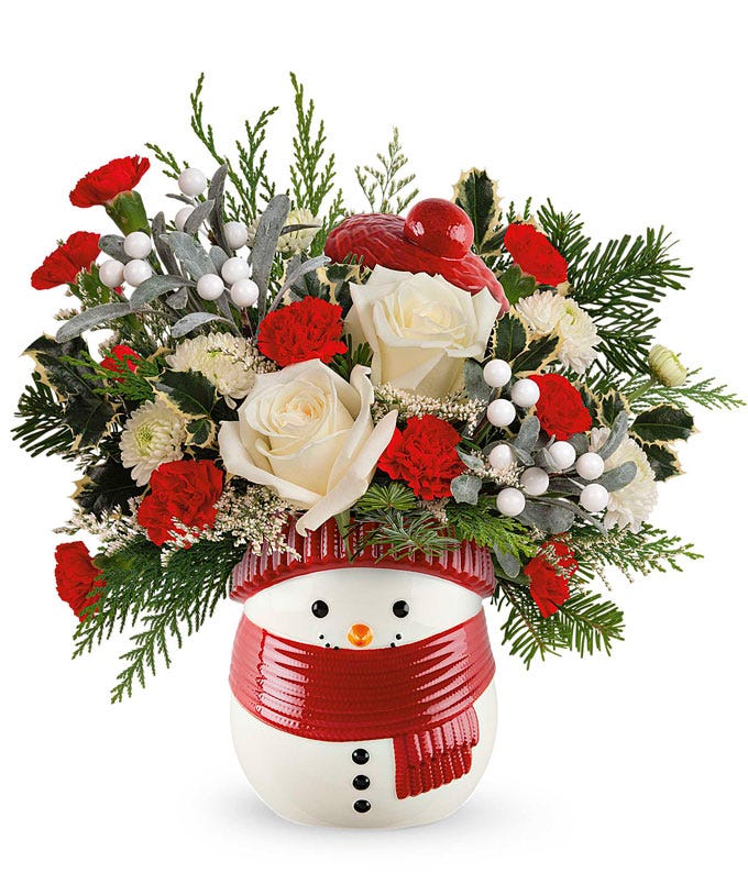 A festive arrangement with white roses, red carnations, miniature carnations, greenery, white berries, and silver leaves, displayed in a snowman container with a red scarf and hat.