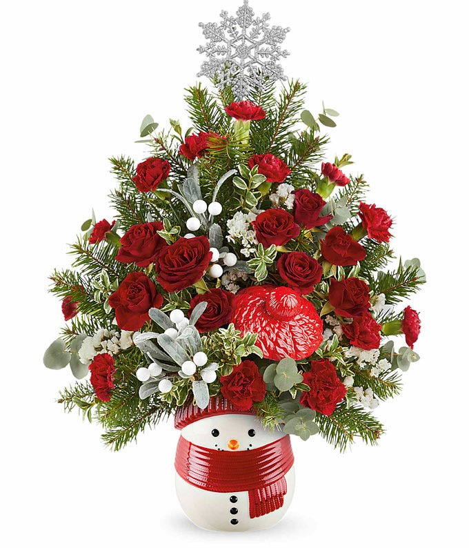 Festive holiday floral arrangement shaped like a Christmas tree, featuring red roses, red carnations, white berry accents, frosted greenery, and a large silver snowflake topper. Presented in a snowman-shaped container wearing a red scarf.