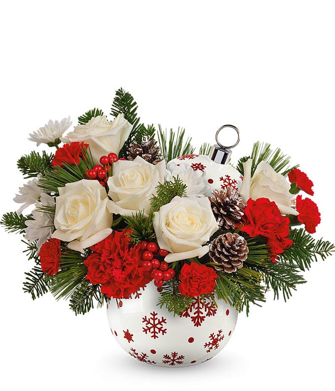 A festive bouquet with white roses, red carnations, pinecones, red berries, and greenery. Arranged in a round ornament-shaped container with red snowflake designs, perfect for holiday decor.