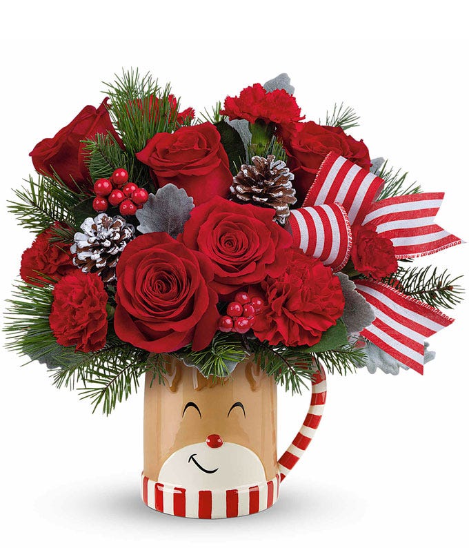 A festive bouquet of red roses, maroon and miniature red carnations, pinecones, red berries, and greenery. Displayed in a smiling reindeer mug with a candy cane handle and striped bow.