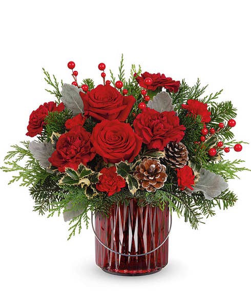 A festive bouquet of red roses, carnations, and miniature carnations with greenery, pinecones, silver leaves, and red berries, arranged in a deep red ribbed glass vase.