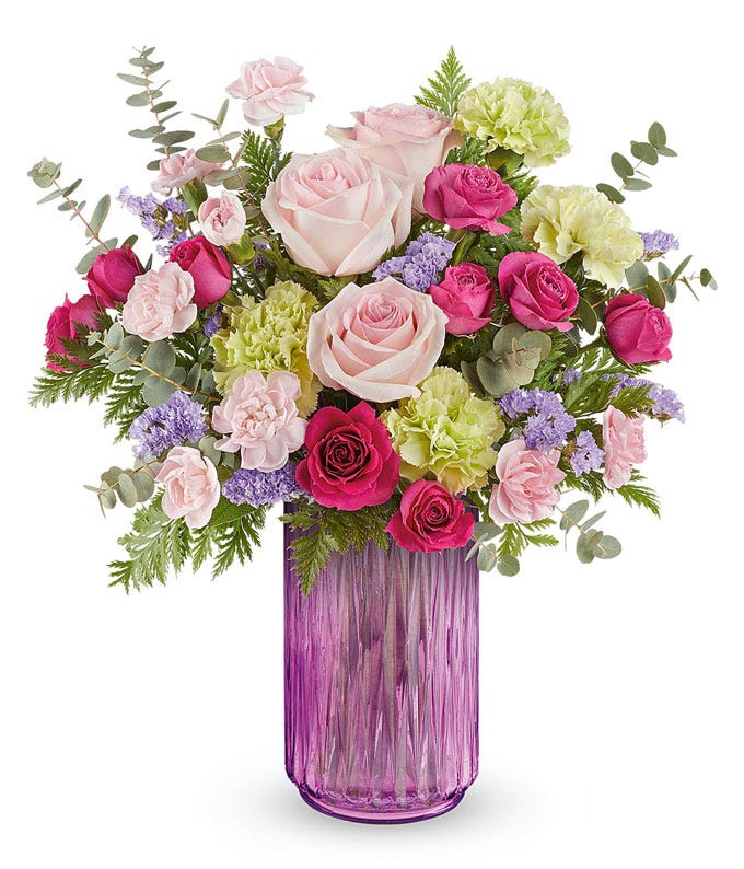 Floral arrangement with peach roses, light pink alstroemeria, miniature peach carnations, lavender daisy spray chrysanthemums, and floral greenery in a textured lavender glass vase.