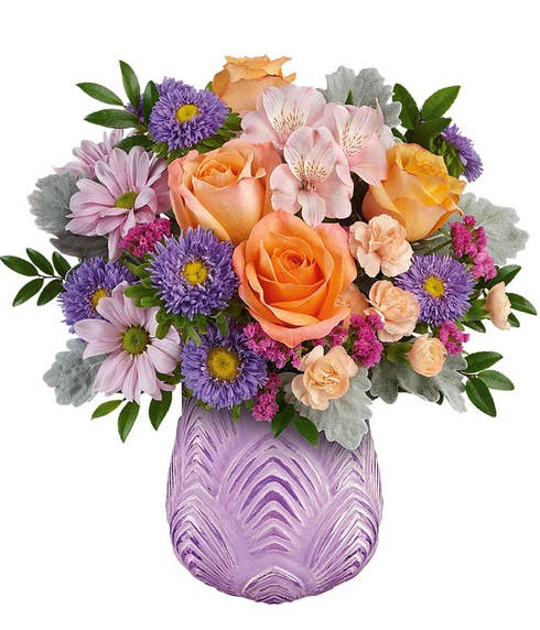 Lush floral arrangement featuring peach roses, pink alstroemeria, miniature peach carnations, lavender daisy spray chrysanthemums, purple aster poms, pink statice, dusty miller, and assorted greenery, elegantly displayed in a textured lavender glass vase.