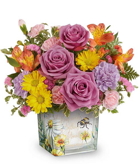 Lavender roses, purple carnations, yellow daisy spray chrysanthemums, orange alstroemeria, pink miniature carnations, and floral greenery, displayed in a decorative keepsake container with a bee and wildflower design.
