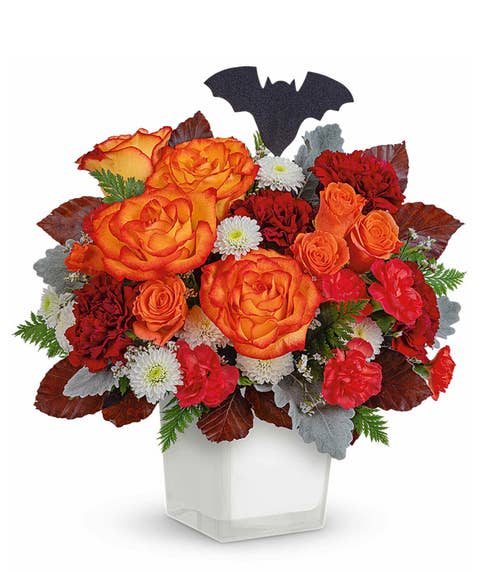 Halloween flower arrangement with orange bi-color roses, red carnations, orange spray roses, and white button chrysanthemums in a white cube vase, accented with fall leaves, greenery, and a black bat decoration.