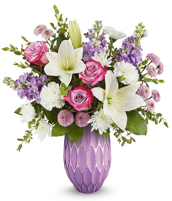 Lavender roses, white asiatic lilies, white carnations, lavender stock, lavender button spray chrysanthemums, white cushion spray chrysanthemums, and floral greenery in a purple vase.