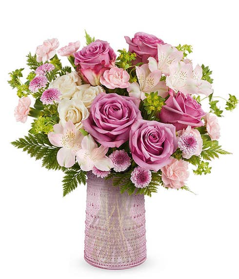 lavender roses, creme spray roses, light pink alstroemeria, miniature pink carnations, purple button spray chrysanthemums, and floral greenery in a pink vase.