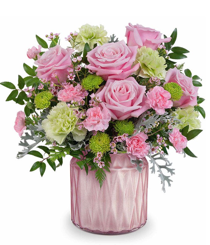 pink roses, green carnations, miniature pink carnations, green button spray chrysanthemums, pink waxflower, and floral greenery in a pink vase.