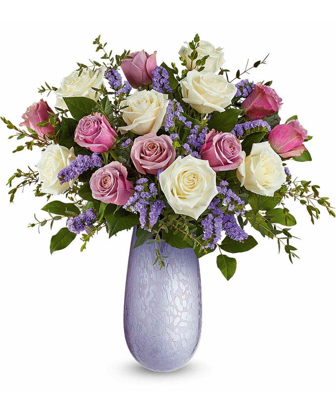 An elegant floral arrangement featuring crème roses, lavender roses, lavender sinuata statice, and fresh floral greenery, arranged in a lavender vase.