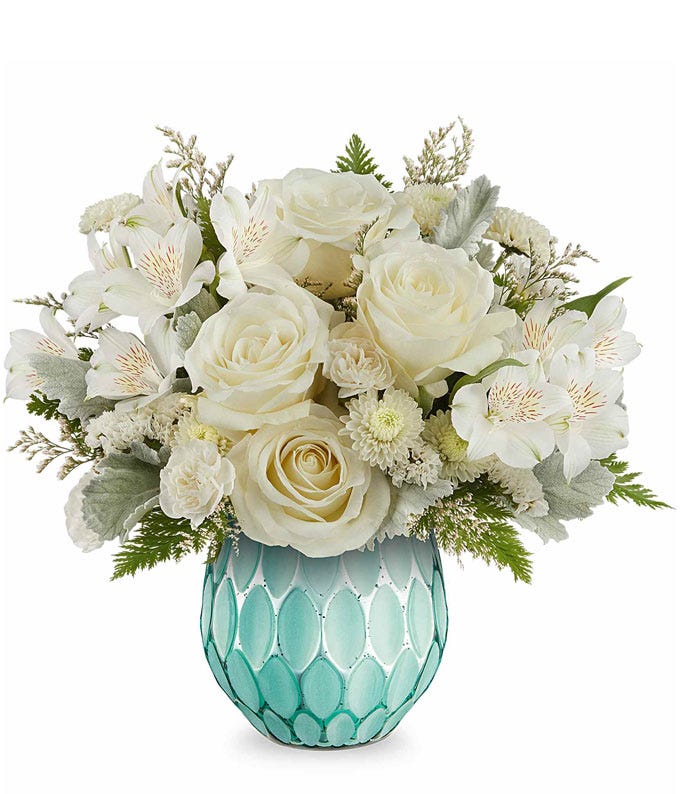 Elegant floral arrangement featuring white roses, white alstroemeria, white button spray chrysanthemums, dusty miller, and assorted greenery, arranged in a textured turquoise glass vase with a geometric pattern.