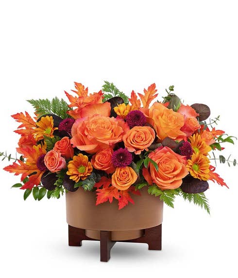 Fall floral arrangement with orange roses, orange spray roses, yellow and red bicolor daisy poms, deep burgundy button spray chrysanthemums, and autumn leaves in a brown ceramic planter with wooden stand.