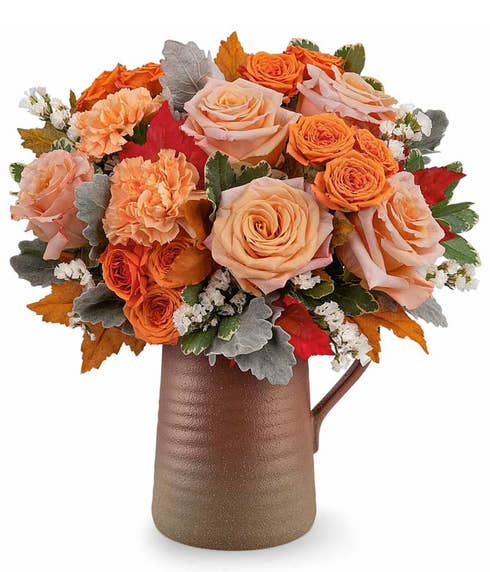 Fall flower arrangement featuring peach and orange roses, orange carnations, white filler flowers, dusty miller, and autumn leaves in a rustic brown ceramic pitcher.