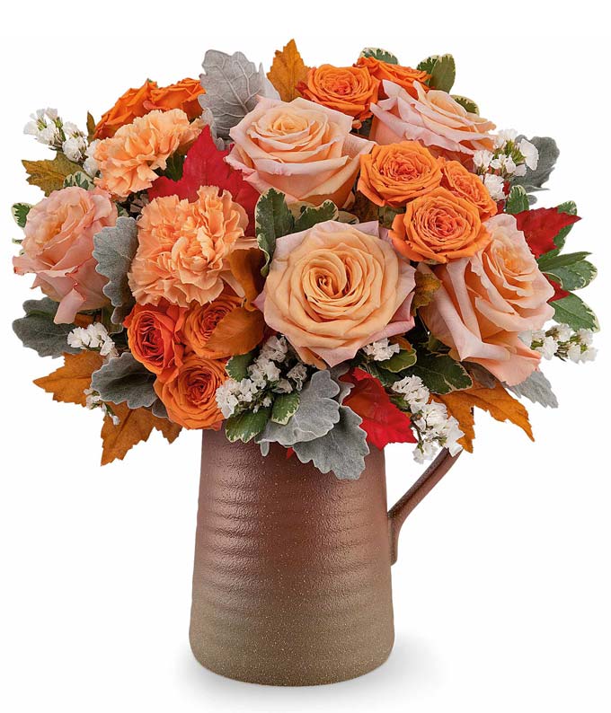 Fall flower arrangement featuring peach and orange roses, orange carnations, white filler flowers, dusty miller, and autumn leaves in a rustic brown ceramic pitcher.