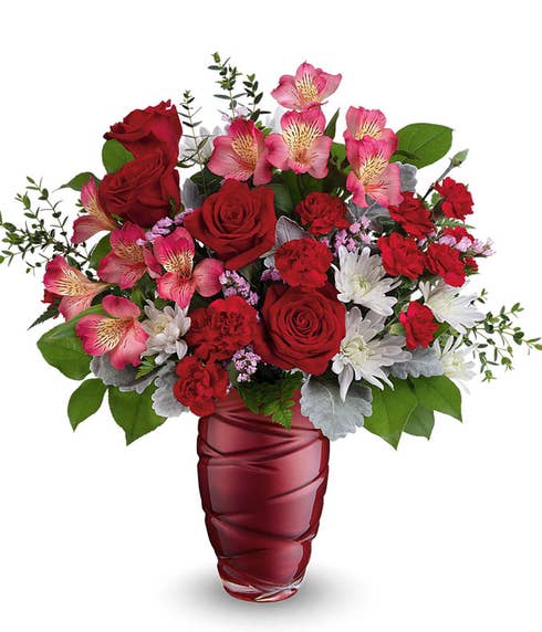 A Valentine's Day bouquet featuring red roses, pink alstroemeria, red carnations, white poms, pink filler flowers, dusty miller, and lush greenery, arranged in a deep red swirl-patterned glass vase.