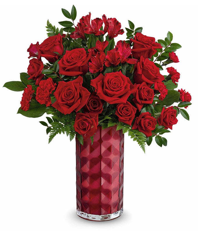 A Valentine's Day bouquet featuring red roses, red spray roses, red alstroemeria, and lush greenery, arranged in a tall faceted red glass vase.
