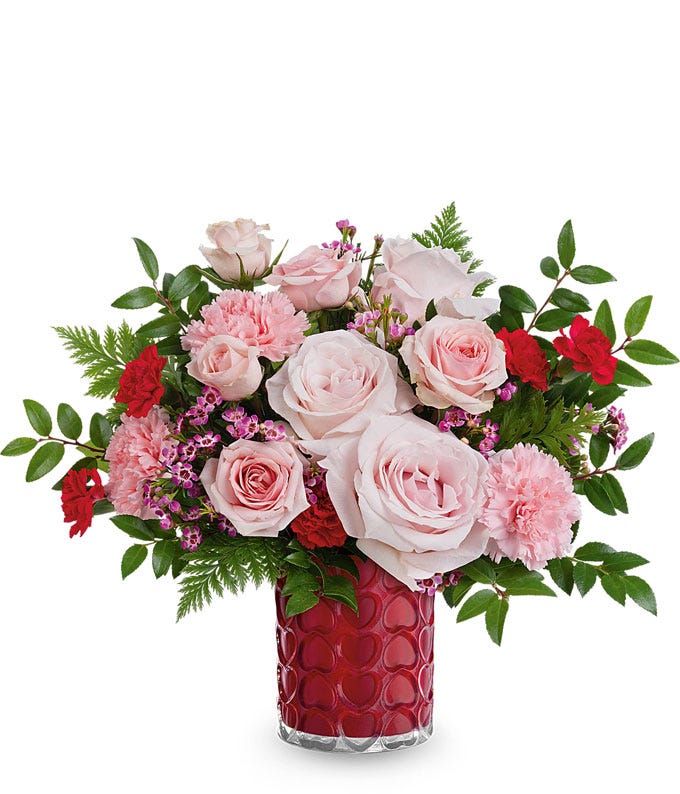 A Valentine's Day bouquet featuring light pink roses, pink carnations, red spray roses, pink waxflower, and lush greenery, arranged in a red glass vase with a textured heart pattern.