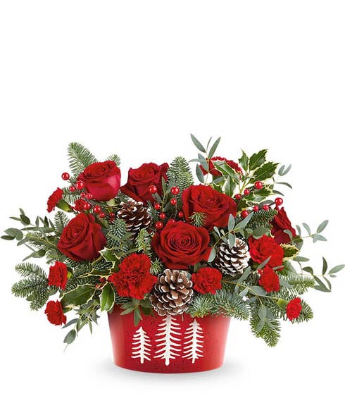 Christmas flower arrangement with red roses, red carnations, and miniature red carnations in a red container with white tree design, accented with pinecones, berries, and evergreen foliage.