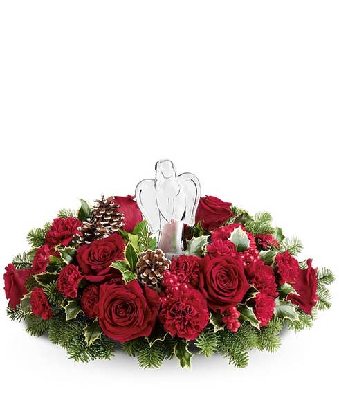 Christmas centerpiece featuring red roses, red carnations, evergreen sprigs, pinecones, holly, red berries, and a central clear glass angel figurine.