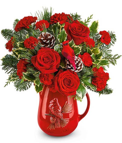 Holiday floral arrangement with red roses, red carnations, miniature red carnations, pinecones, holly, greenery, and a cardinal pick in a red ceramic pitcher featuring a raised cardinal design.