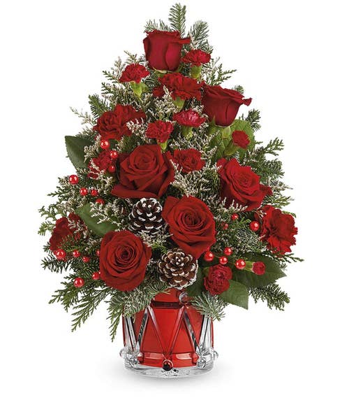 Christmas tree-shaped floral arrangement with red roses, red mini carnations, white limonium, pinecones, red berry spray, and greenery in a red drum-shaped container.