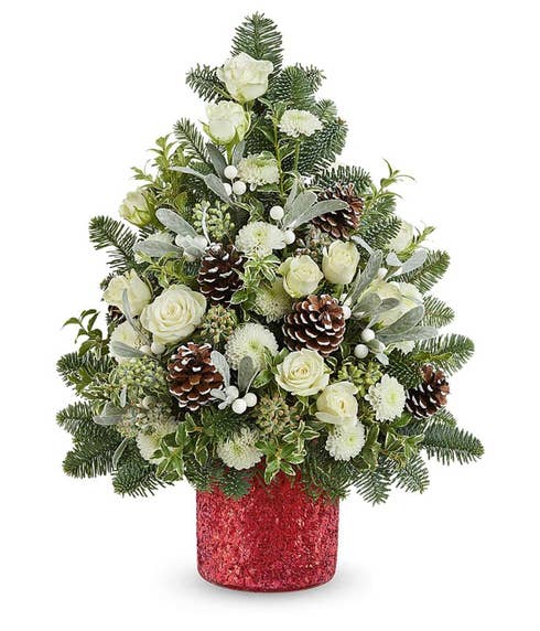 Christmas tree-shaped floral arrangement with white roses, white carnations, frosted pinecones, white berry spray, dusty miller, and seasonal greenery in a red glittered container.