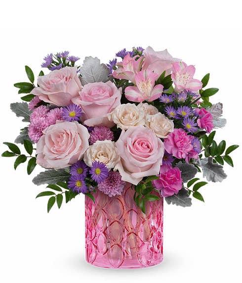 Pink Roses, Creme Spray Roses, Pink Alstroemeria, Mini Hot Pink Carnations, Lavender Button poms, and floral greenery in a pink textured glass cylinder vase