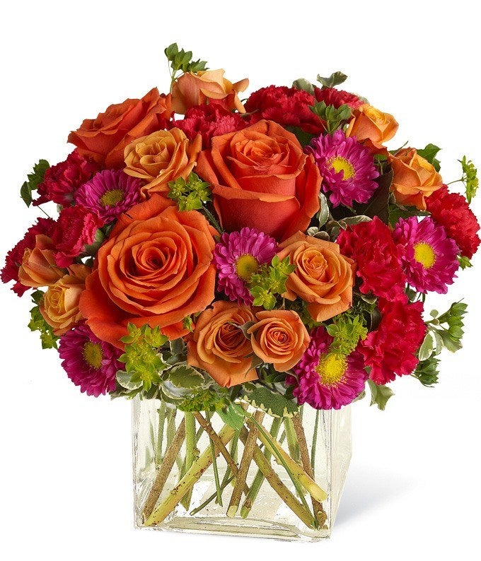 A combination of light orange roses and orange spray roses with hot pink carnations and Matsumoto asters on a clear vase
