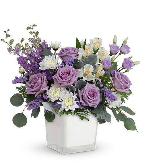 Lavender roses, white alstroemeria, lavender lisianthus and larkspur, white mums, lavender statice , and floral greens in a white glass cube vase