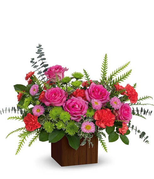 Hot pink roses, orange carnations and mini carnations, pink asters, green chrysanthemums, spiral eucalyptus, and floral greens in a brown bamboo cube vase 