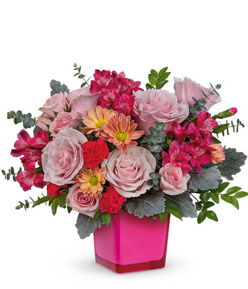Pink roses, dark pink alstroemeria, hot pink carnations, eucalyptus, and floral greens in a neon pink glass cube vase
