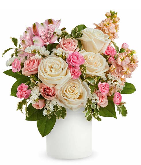 A Mother's Day arrangement featuring crme roses, pink spray roses, pink alstroemeria, miniature pink carnations, peach stock, and floral greens, arranged in a white cylinder vase, with space for a personalized gift message.