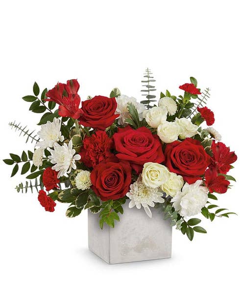 Red roses with white roses, white poms, red carnations, and lush greenery arranged in a modern white cube vase