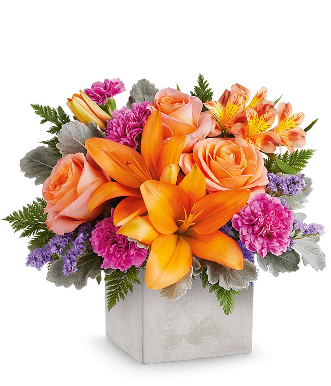 Orange roses, orange Asiatic lilies, orange alstroemeria, fuchsia carnations, and floral greenery in a modern cube vase with a faux concrete look