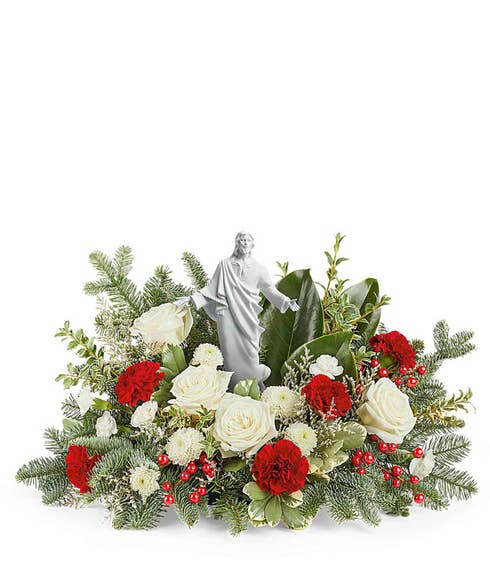 Floral arrangement featuring white roses, red carnations, miniature white carnations, and seasonal greenery. Accented with red berries and a serene angel figurine at the center.