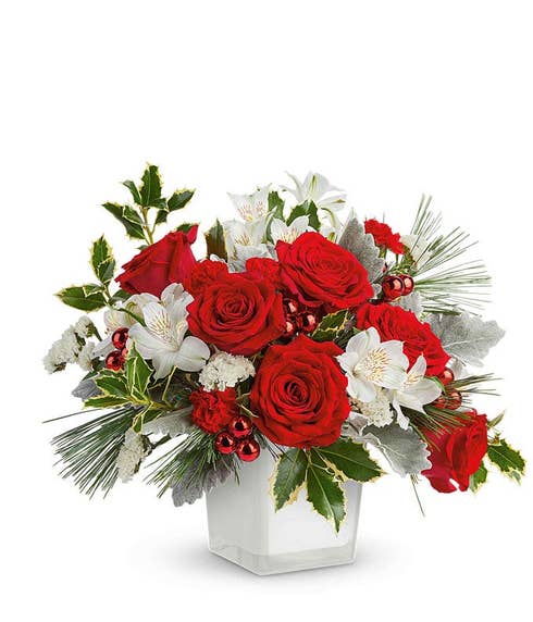 Christmas floral arrangement with red roses, white alstroemeria, white carnations, red berry ornaments, pine greenery, silver dusty miller, and holly in a white cube container.