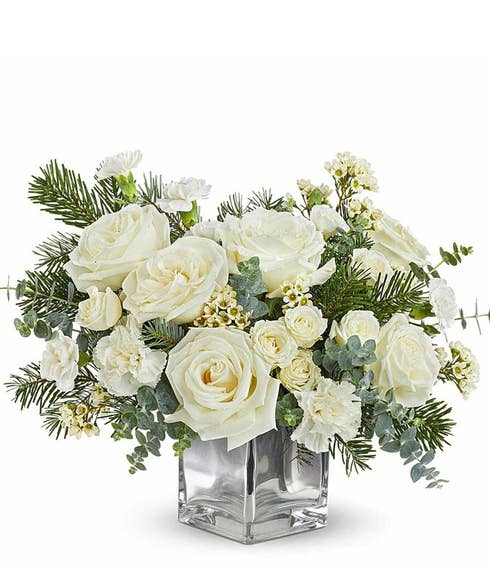White roses, spray roses, miniature carnations, and waxflower arranged with greenery and evergreens in a silver cube vase.